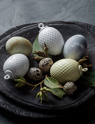 Rhombe Ostereier aus Porzellan in den Farben Pastellgelb und Weiß von Lyngby Porcelæn, arrangiert mit weiteren Eiern aus Porzellan, Wachteleiern und grünen Zweigen auf einer schwarzen Schieferplatte