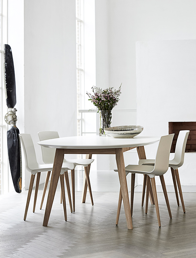 Andersen Furniture DK10 Esstisch mit weißer HPL-Tischplatte und Beinen aus Eichenholz, kombiniert mit weißen Esszimmerstühlen in einem hellen Wohnraum.
