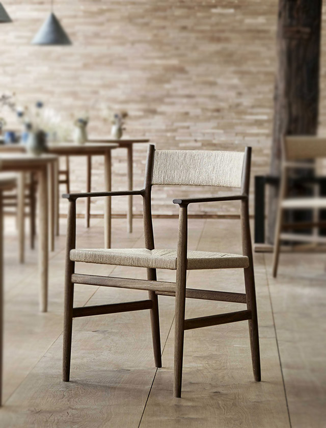 Das Bild zeigt den Arv Chair des dänischen Herstellers Brdr. Krüger. Der Stuhl ist aus geräucherter, geölter Eiche gefertigt und besitzt eine handgeflochtene Sitzfläche aus Naturgeflecht (Papierkordel). Mit seiner klaren Linienführung und dem reduzierten skandinavischen Design verbindet er traditionelle Handwerkskunst mit moderner Eleganz.