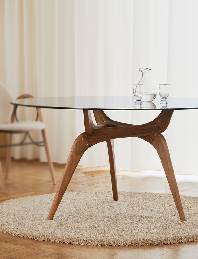 Der Triiio Esstisch von Brdr. Krüger kombiniert ein organisch geformtes Gestell aus Räuchereiche mit einer eleganten Tischplatte aus Bronzeglas und steht in einem hellen, wohnlichen Ambiente.