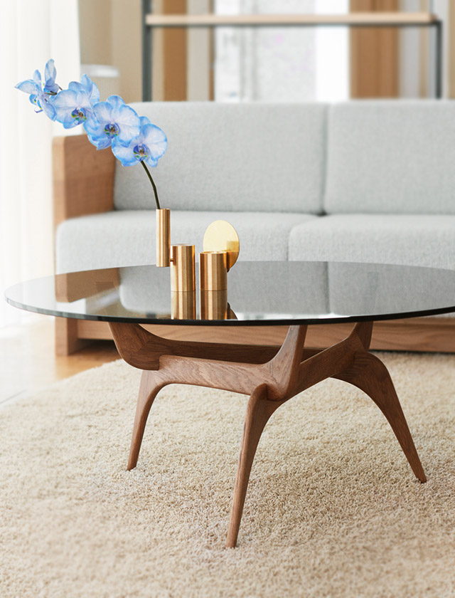 Der Triiio Couchtisch von Brdr. Krüger kombiniert ein organisch geformtes Gestell aus Räuchereiche mit einer eleganten Tischplatte aus Bronzeglas und steht vor dem Karm Sofa in einem hellen, wohnlichen Ambiente.