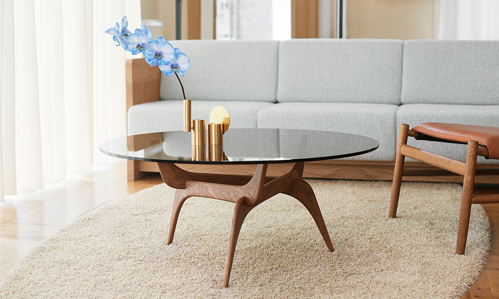 Der Triiio Couchtisch von Brdr. Krüger kombiniert ein organisch geformtes Gestell aus Räuchereiche mit einer eleganten Tischplatte aus Bronzeglas und steht vor dem Karm Sofa in einem hellen, wohnlichen Ambiente.