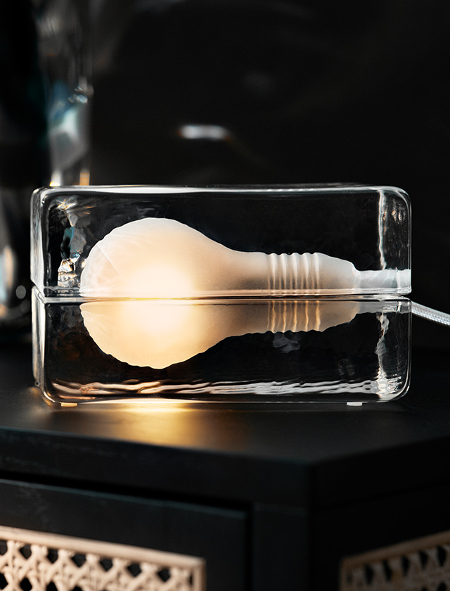 Design-Tischlampe im Stil einer eingeschlossenen Glühbirne, die in einem klaren Glasblock leuchtet. Moderne und minimalistische Beleuchtung mit warmem Licht, platziert auf einem dunklen Möbelstück.