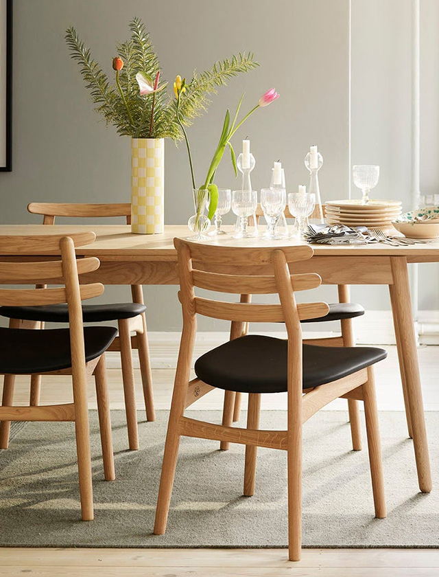 Andersen Furniture Mood Chair Armlehnstühle, die sich an einem dunklen, ovalen Tisch gegenüberstehen. Die Stühle kombinieren ein helles Holzgestell mit weich gepolsterten Sitz- und Rückenflächen in einem warmen, beigefarbenen Kvadrat-Stoff. Auf dem Tisch liegen ein aufgeschlagenes Magazin, einige Bücher sowie ein Stift, wodurch eine einladende, wohnliche Atmosphäre entsteht. Die Aufnahme aus der Vogelperspektive betont die klare Formensprache, das minimalistische skandinavische Design und die harmonische Verbindung von natürlichen Materialien mit modernen Linien.