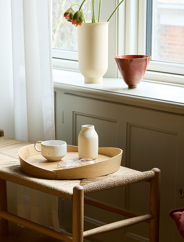 Ruhige, wohnliche Szene mit dem V43 Tablett aus FSC®-zertifiziertem Eichenholz, das auf einer Sitzbank platziert ist. Auf dem Tablett befinden sich eine Keramiktasse, eine kleine Vase und eine Lesebrille, was auf eine entspannte Kaffee- oder Lesepause hinweist. Im Hintergrund sind ein Fenster, durch das natürliches Licht einfällt, und zwei Keramikgefäße auf der Fensterbank zu sehen, eines davon mit welkenden Blumen. Diese Anordnung unterstreicht die Eleganz und Funktionalität des Tabletts, das sich nahtlos in unterschiedliche Einrichtungsstile einfügt und für den täglichen Gebrauch geeignet ist.