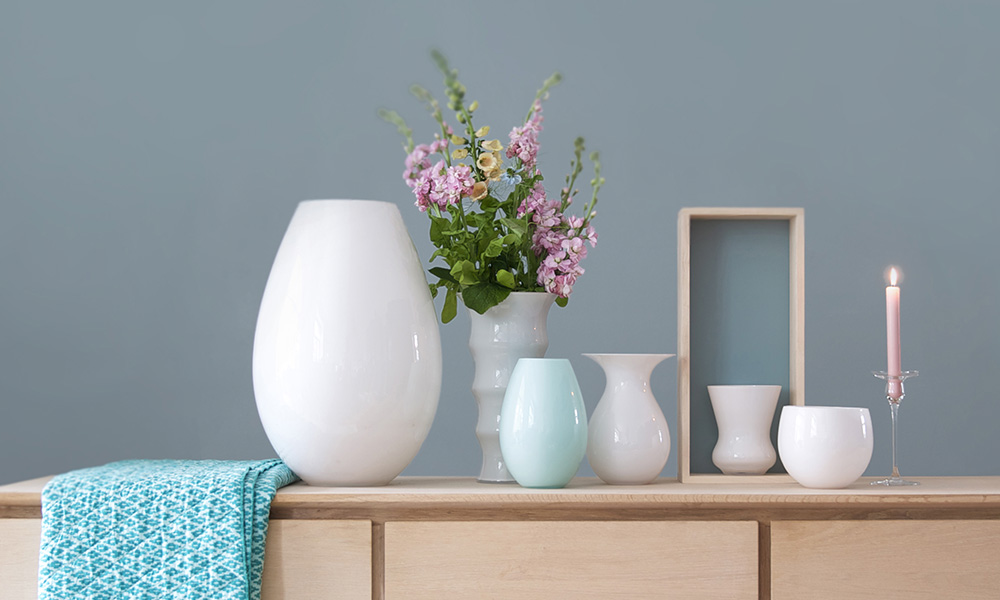 Das Bild zeigt Glasvasen aus mundgeblasenem Glas von Holmegaard aus Dänemark, aufgestellt auf einem Sideboard vor einer grauen Wand. Ein besonderer Blickfang ist die weiße Cocoon-Bodenvase. Weiterhin zu sehen sind eine kleine Cocoon-Vase aus hellblauem Glas, ein Cocoon-Übertopf, die Karen Blixen - Glasvase mit einem Blumenstrauß bunter Sommerblumen. Die Karen-Blixen-Vase wurde von Anja Kjaer, die Shape-Glasvase und die Cocoon-Vasen von Peter Svarrer entworfen.