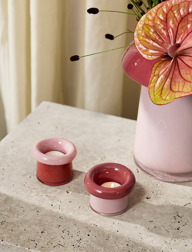 Holmegaard Soma Teelichthalter Rose/Terrakotta – mundgeblasenes Glas auf einer Tischplatte aus Stein stehend mit jeweils einem angezündetem Teelicht, daneben steht die Soma Vase in Rosé/ Terracotta mit einem Blumenbouquet