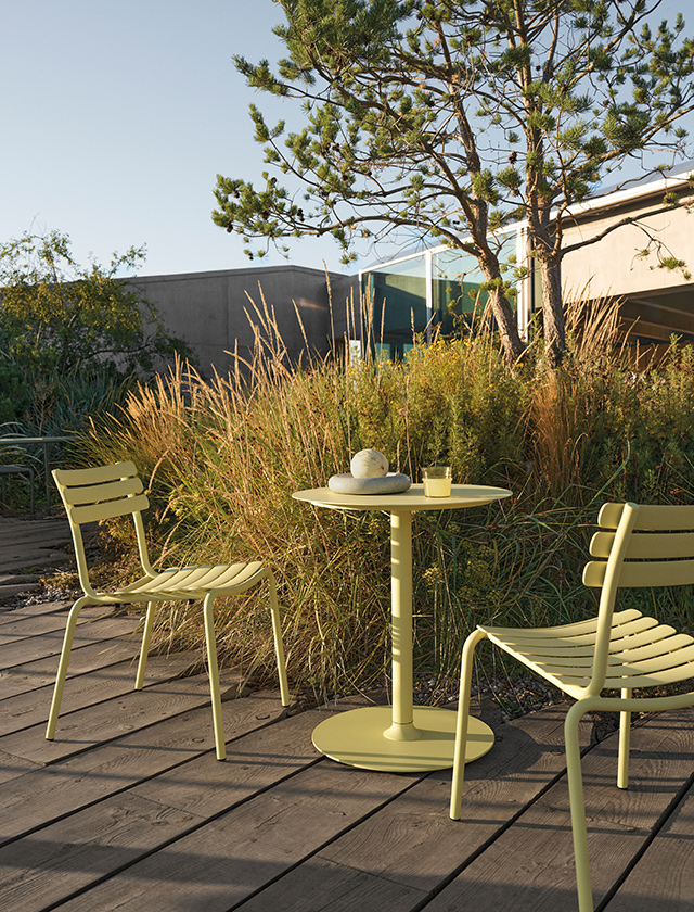 Zwei HOUE ALUA Gartenstühle aus Aluminium in Lemon Zest mit passendem PICO Bistrotisch auf einer modernen Holzterrasse. Das stilvolle Outdoor-Set eignet sich ideal für Garten, Terrasse, Balkon sowie den Einsatz im Café- oder Restaurant-Außenbereich.