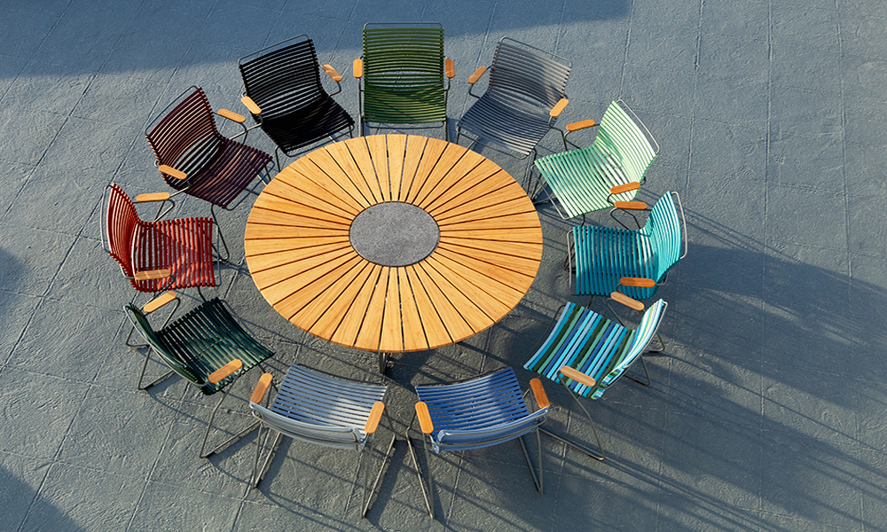 HOUE Click Dining Chairs in den Farben Pine green, Paprika, Black, Plum, Grey, Dark grey, Olive green, Dusty light green, Petrol, Dusty light blue, Pigeon blue und Dark blue in einem Kreis aufgestellt. In der Mitte steht der Circle Table mit Lamellen aus Bambus und einer im Zentrum befindlichen Steinplatte zum Abstellen von Heißen Topfen und Tischgrills.