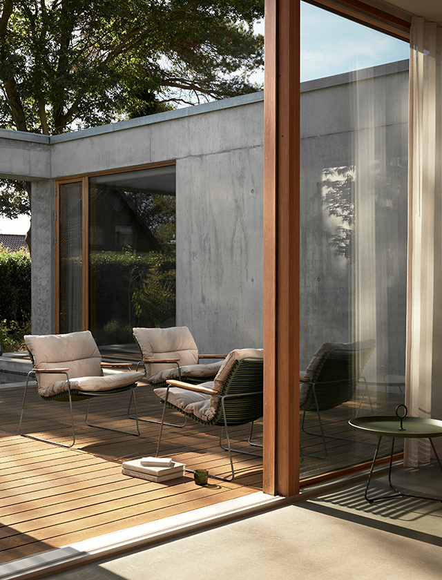 Stilvolle Outdoor-Szene auf einer modernen Terrasse mit dem HOUE Click Roomy Chair Assemble und passenden HOUE Click Sitzkissen. Die Lounge-Stühle mit filigranem Metallgestell, charakteristischen Kunststofflamellen und hellen Polstern stehen auf einem Holzdeck vor puristischer Beton- und Glasarchitektur und schaffen eine elegante, wohnliche Atmosphäre im Außenbereich.