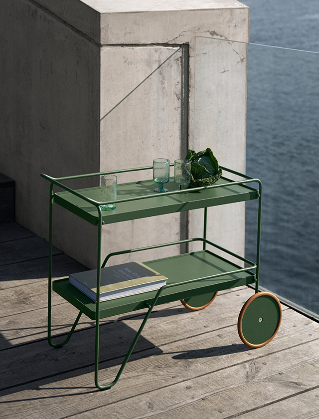 Der HOUE CLICK Servierwagen in Olive Green ist ein minimalistischer Outdoor-Servierwagen mit zwei Ablagen und Rollen. Im Bild steht der Wagen auf einer Holzterrasse am Wasser und ist mit Gläsern und einem Buch dekoriert.