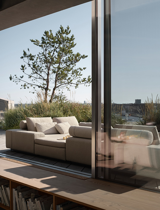 Modernes HOUE DRIFT Outdoor-Lounge-Sofa in heller Stoffoptik auf einer sonnigen Terrasse mit Gräsern, Baum und großzügiger Fensterfront.