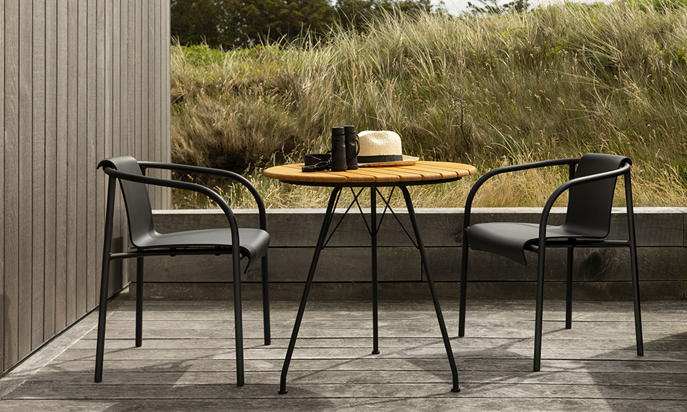 Moderne Terrasse in Jütland, Dänemark, stilvoll möbliert mit hochwertigem Outdoor-Design der Marke HOUE. Im Mittelpunkt stehen zwei elegante NAMI Dining-Stühle mit Armlehne in Schwarz, deren geschwungenes, organisches Design aus recyceltem Kunststoff und pulverbeschichtetem Metallrahmen typisch für das skandinavische Stilgefühl ist. Dazwischen befindet sich ein runder CIRCUM Gartentisch mit einer edlen Bambus-Tischplatte und schlankem, schwarzem Stahlgestell.