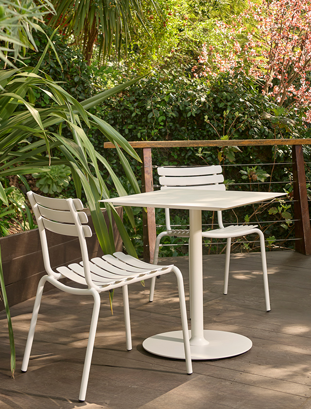 Outdoor-Sitzbereich mit einem quadratischen HOUE PICO Gartentisch (60×60?cm) in der dezenten Farbe Muted White. Um den Tisch stehen zwei passende ALUA Dining Stühle ohne Armlehne, ebenfalls in Muted White. Die Szene ist eingebettet in eine üppige, grüne Gartenterrasse mit tropischem Flair und natürlichem Lichteinfall. Diese hochwertige und wetterfeste Möbelkombination vereint minimalistisches Design mit funktionaler Outdoor-Nutzung – ideal für Balkon, Terrasse oder Garten.