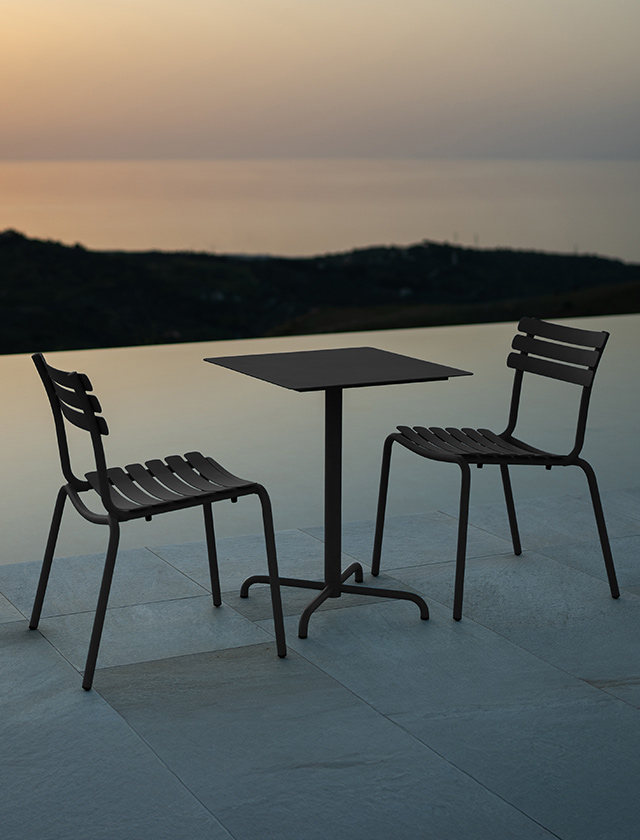 Outdoor-Szene bei Sonnenuntergang mit einem quadratischen HOUE PICO Gartentisch in Schwarz (60×60?cm) und zwei dazu passenden ALUA Dining Stühlen ohne Armlehnen, ebenfalls in Schwarz. Die minimalistische Möbelkombination steht auf einer modernen Terrasse mit Blick auf das Meer oder einen ruhigen See. Die Atmosphäre ist ruhig und elegant – perfekt für stilvolle Abende im Freien. Ideal für den Einsatz auf Terrassen, Balkonen oder in modernen Gartenanlagen.