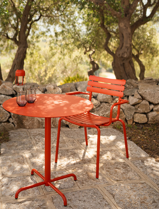 Outdoor-Sitzplatz mit dem runden Gartentisch HOUE PICO in der kräftigen Farbe Cayenne-Rot, kombiniert mit einem passenden Alua-Stuhl im selben Farbton. Die Möbel stehen auf einer Naturstein-Terrasse, eingerahmt von einer rustikalen Trockensteinmauer und mediterranen Olivenbäumen im Hintergrund. Zwei elegante Gläser auf dem Tisch unterstreichen die einladende Atmosphäre. Die Szene vermittelt modernes skandinavisches Design, Langlebigkeit und perfekte Eignung für Garten, Terrasse oder Balkon.