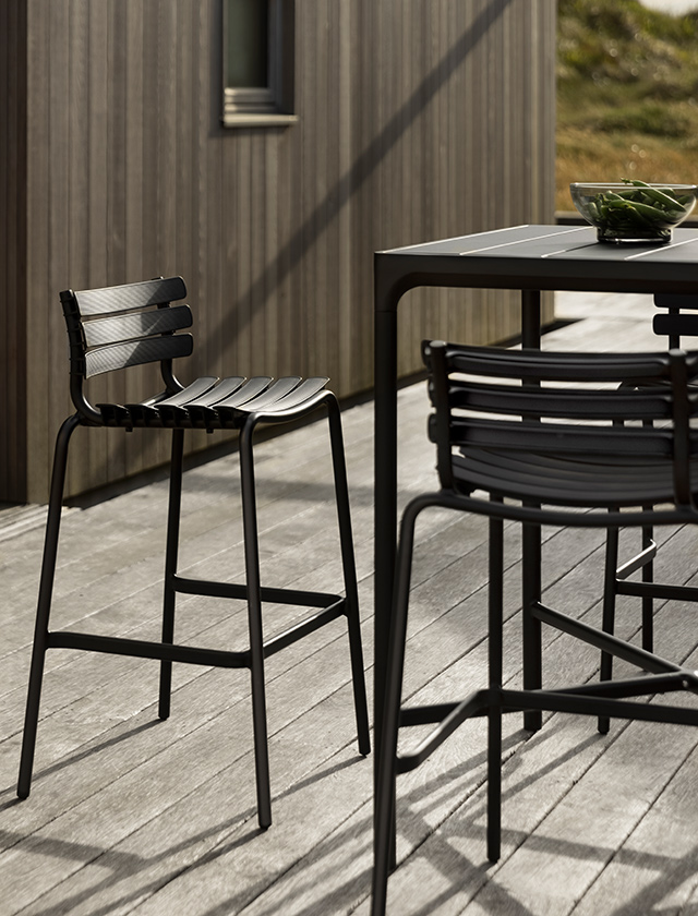 Stilvollen Außenbereich mit modernen Gartenmöbeln von HOUE: ReCLIPS Barhocker in Schwarz und einen dazu passenden FOUR Bartisch. Das Ensemble steht auf einer Holzterrasse, deren Dielen ein warmes Ambiente kreieren, das durch das Spiel von Licht und Schatten noch unterstrichen wird. Auf dem Tisch befindet sich eine Schale mit grünen Pflanzen, was einen Hauch von Natur hinzufügt und die schlichte Eleganz der Möbelstücke ergänzt.