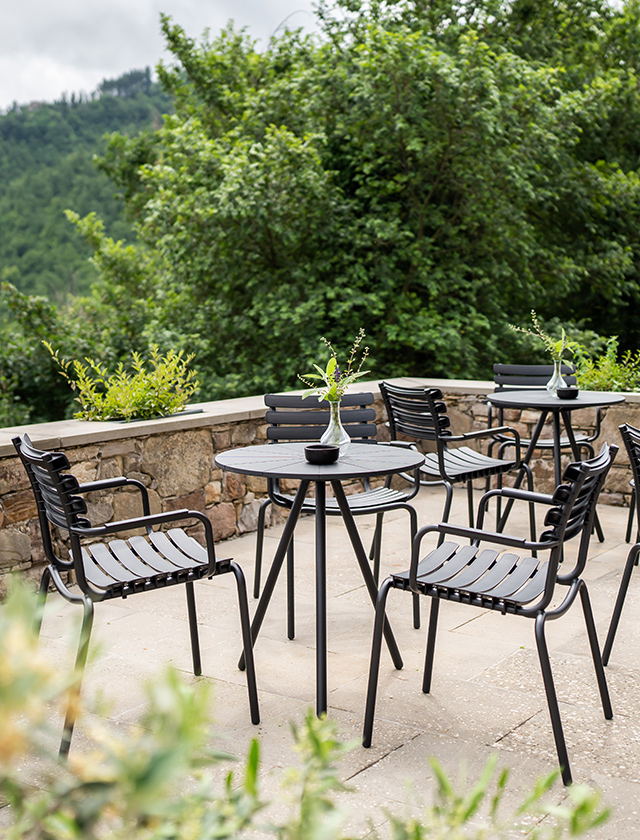 Gemütlicher Außenbereich auf einer Terrasse eines Cafés. Im Vordergrund sieht man mehrere runde Nami Bistrotische von HOUE, die mit schwarzen ReClips Gartenstühlen mit Armlehnen umgeben sind. Auf jedem Tisch steht eine kleine schwarze Vase mit einer zarten grünen Pflanze, was für ein ansprechendes, minimalistisches Detail sorgt. Im Hintergrund erkennt man üppiges Grün von Bäumen und Sträuchern, was darauf hindeutet, dass der Bereich in einer grünen, hügeligen Gegend liegt. Das Ambiente wirkt friedlich und einladend, ideal für eine entspannende Pause im Freien.