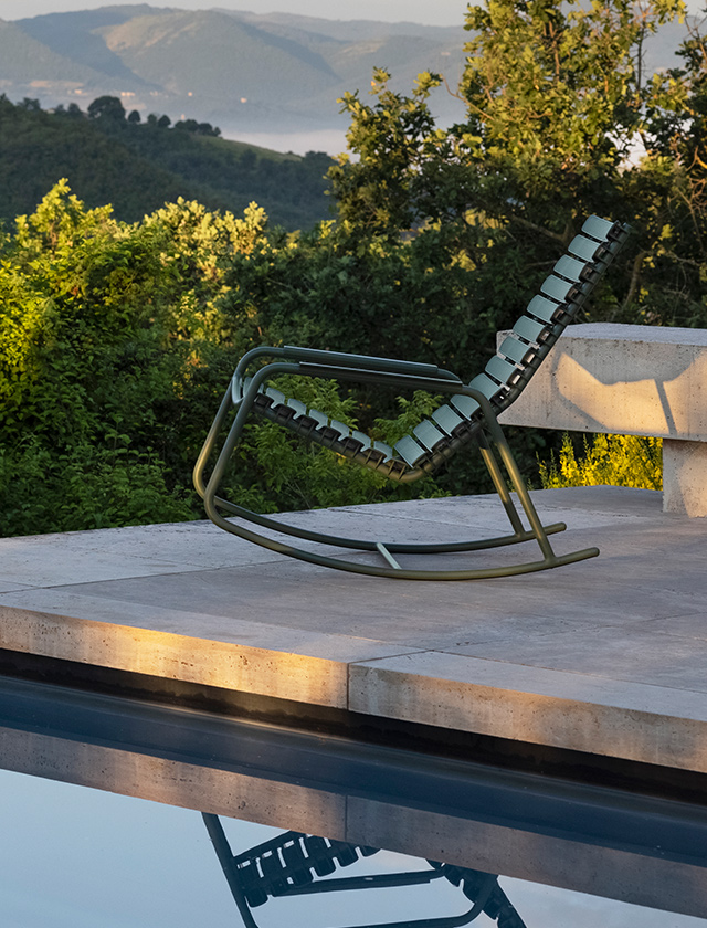 Das Foto zeigt einen olivgrünen "ReCLIPS" Schaukelstuhl von HOUE am Rande eines Infinity-Pools, der eine beruhigende Symmetrie durch die Spiegelung im Wasser erzeugt. Die Szene wird durch die frühe oder späte Tageszeit beleuchtet, die dem Bild eine warme Atmosphäre verleiht. Im Hintergrund erstrecken sich malerische Hügel, die mit Bäumen bedeckt sind und von Nebel umhüllt werden, was eine ruhige, fast mystische Stimmung schafft. Der Schaukelstuhl scheint ein idealer Ort zu sein, um in dieser friedlichen Umgebung zu entspannen und die atemberaubende Aussicht zu genießen.