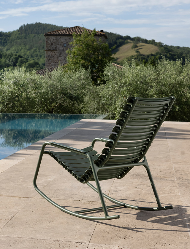 Olivgrüner ReCLIPS Schaukelstuhl von HOUE, der auf einer gepflasterten Terrasse neben einem Infinity-Pool steht. Die Umgebung wirkt friedlich und malerisch mit einer traditionellen Steinmauer und einem alten Steinturm im Hintergrund, umgeben von sanften Hügeln und üppigem Grün. Die Szenerie vermittelt ein Gefühl von Ruhe und Entspannung in der ländlichen Landschaft, und der Schaukelstuhl selbst lädt zum Verweilen und Genießen der natürlichen Schönheit ein.
