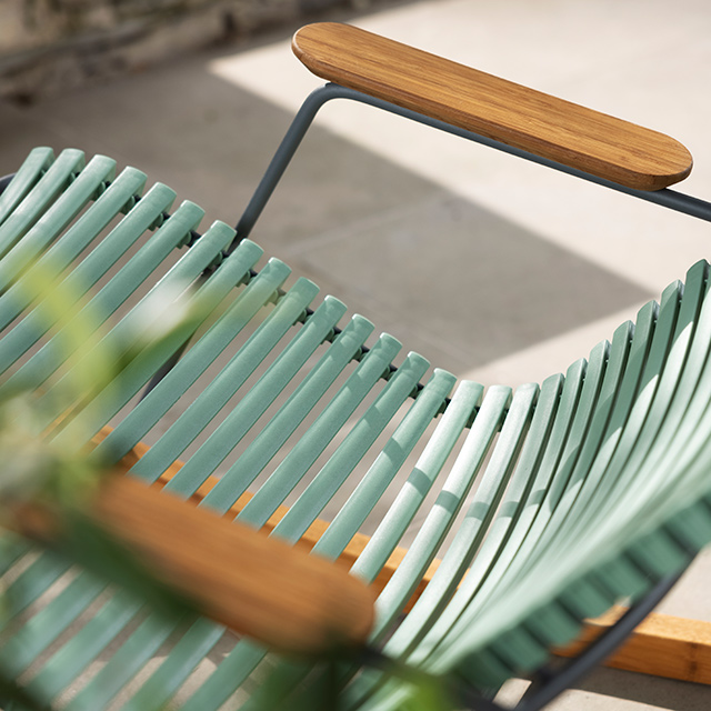 Diese Detailaufnahme zeigt den eleganten HOUE CLICK Rocking Chair in der Farbvariante dusty light green (staubiges Hellgrün). Die sanft geschwungenen Lamellen der Sitzfläche aus wetterfestem Kunststoff schmiegen sich ergonomisch an den Körper an und verleihen dem Schaukelstuhl höchsten Sitzkomfort. Die Armlehnen aus hochwertigem Bambusholz setzen einen natürlichen Kontrast zur modernen Linienführung des Gestells. Das minimalistische, skandinavische Design eignet sich ideal für den Einsatz auf Balkon, Terrasse oder im Garten und verbindet Funktionalität mit stilvoller Ästhetik.