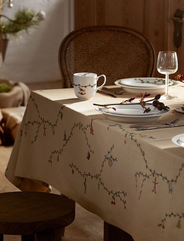 Elegante Kähler Hammershøi Christmas Tischdecke in Sandfarbe zu sehen. Das 150x220 cm große Tafeltuch bildet die stilvolle Basis eines weihnachtlich gedeckten Tisches. Entlang der Ränder sind dezente Weihnachtsmotive mit fein verzierten Zederzweigen und kleinen Lärchenzapfen zu erkennen – ein diskretes Design, das auch beim Servieren mit Schüsseln und Tellern sichtbar bleibt.