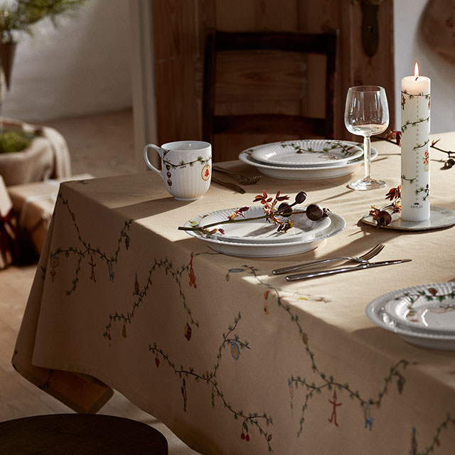 Kähler Hammershøi Christmas Tischdecke in sanftem Sandton, 150x220 cm. Das feine Baumwollgewebe ziert einen festlich gedeckten Tisch und zeigt an den Rändern zurückhaltende Weihnachtsillustrationen mit geschmückten Zweigen und kleinen Zapfen. Das dezente Muster bleibt auch unter Geschirr und Servierschalen erkennbar und sorgt für eine elegante, saisonale Atmosphäre.