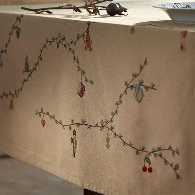 Elegante sandfarbe Kähler Hammershøi Christmas Tischdecke. Entlang der Ränder schmücken feine weihnachtliche Ornamente mit Tannenzweigen und kleinen Zapfen das Gewebe. Das zurückhaltende Design bleibt auch beim Eindecken mit Tellern und Schüsseln sichtbar und verleiht dem Tisch eine festliche wie zugleich stilvolle Ausstrahlung.“