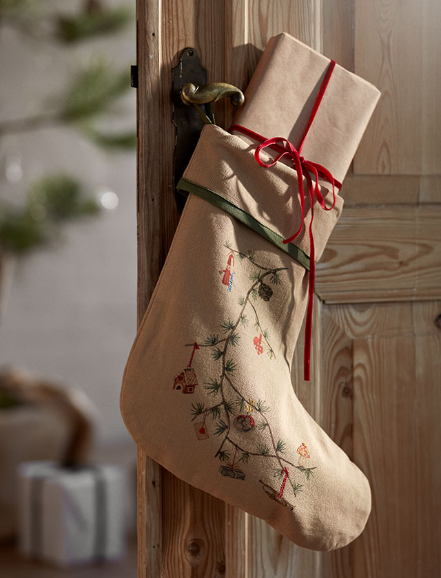 Kähler Hammershøi Christmas Weihnachtsstrumpf an einer Tür, gefüllt mit einem Geschenk. Der 46 cm lange Strumpf ist aus festem Canvas gefertigt und zeigt einen dekorativen Zederzweig mit kleinen Weihnachtsmotiven, die von Rikke Jacobsen illustriert wurden. Ein dunkelgrünes Einfassband am Umschlagrand setzt einen eleganten Akzent und verbindet den Strumpf harmonisch mit dem Hammershøi Christmas Baumteppich und dem restlichen Sortiment. Nachhaltig aus 82 % recycelter Baumwolle und 18 % Polyester hergestellt und GOTS-zertifiziert, vereint er Qualität, Funktionalität und Design. Der Weihnachtsstrumpf ist Teil der beliebten Hammershøi Christmas Serie von Kähler, die klassische Tradition und modernes skandinavisches Design stilvoll verbindet.