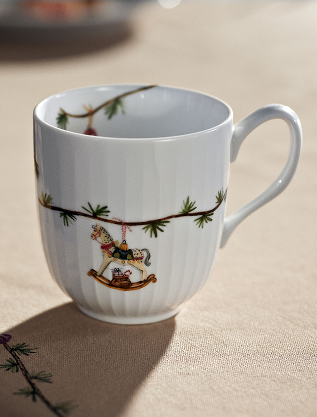 Kähler Design Hammershøi Weihnachtstasse 2025 aus feinem weißen Porzellan, verziert mit Tannenzweigen und detailreichem Schaukelpferd-Motiv, stilvoll platziert auf einem beigen Hammershøi-Tischtuch – skandinavisches Weihnachtsdesign in eleganter Ausführung.