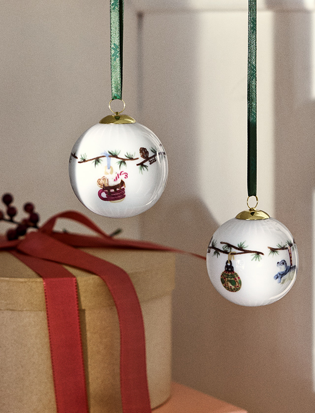 Kähler Design Hammershøi Jul Christbaumkugeln 2026 mit liebevoll illustrierten Weihnachtsmotiven und grünen Bändern, dekoriert über einem festlich verpackten Geschenk.