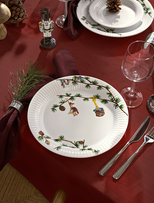 Kähler Design Hammershøi Jul Weihnachtsteller 2026 mit Tannenzweigen und liebevollen Weihnachtsmotiven auf einer festlich gedeckten Tafel in Rot.