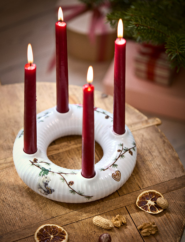 Kähler Design Hammershøi Jul Adventskranz aus weißem Porzellan mit vier roten Kerzen, Tannenzweigen und klassischen Weihnachtsmotiven als festliche Adventsdekoration auf einem Holztisch.