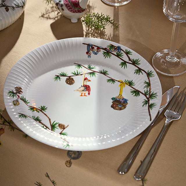 Kähler Design Hammershøi Jul Weihnachtsteller 2027 aus weißem Porzellan mit Tannenzweigen, Zapfen und liebevoll illustrierten Weihnachtsmotiven auf festlich gedecktem Tisch.