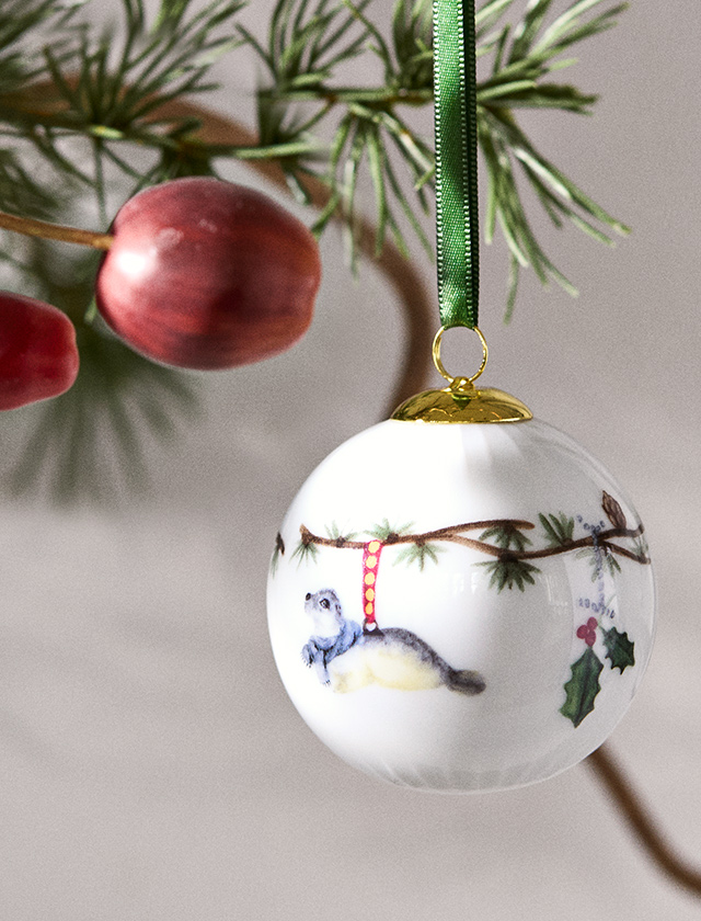 Kähler Design Hammershøi Jul Christbaumkugel 2026 aus weißem Porzellan mit Tannenzweigen, farbigem Tiermotiv, goldfarbener Aufhängung und grünem Band.