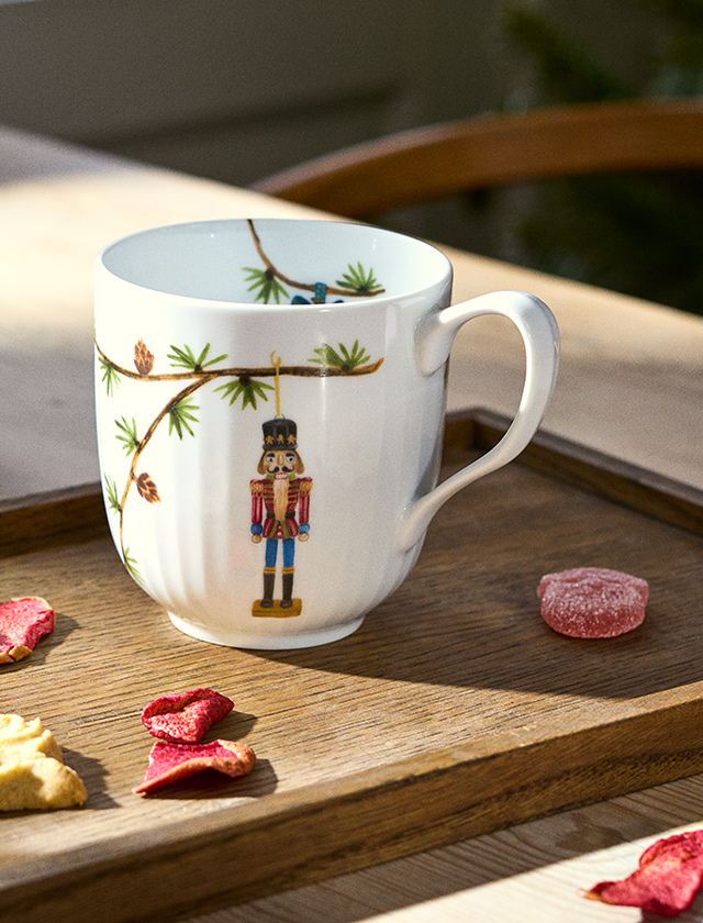 Kähler Design Hammershøi Jul Weihnachtstasse 2026 aus weißem Porzellan mit Nussknacker, Tannenzweigen und Zapfen, dekoriert auf einem Holztablett mit weihnachtlichen Süßigkeiten.