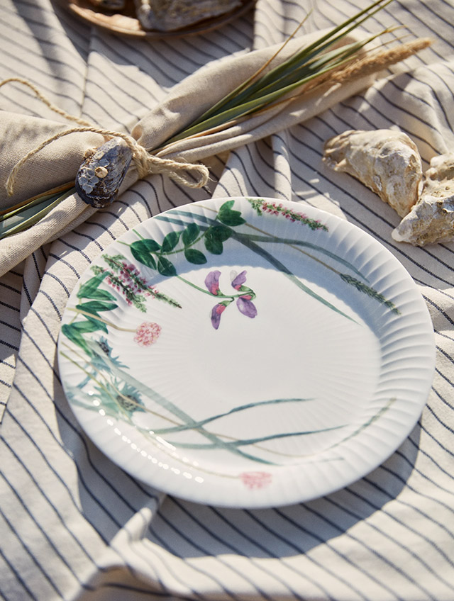 Blühende Küstenpflanzen, Muscheln und kleine Strandtiere – in sanften, gedeckten Farben auf weißem Porzellan. Die Serie Hammershøi Summer 2026 von Kähler bringt die stille Poesie eines Spaziergangs an der dänischen Küste auf den Tisch. Die feinen Aquarellmotive der Künstlerin Rikke Jacobsen scheinen dabei über die charakteristischen Rillen des von Hans-Christian Bauer entworfenen Geschirrs zu wachsen. Ein Teller für das sommerliche Frühstück, das kalte Buffet oder den Kaffee im Freien – und eine stimmige Ergänzung zu den übrigen Stücken der Serie.