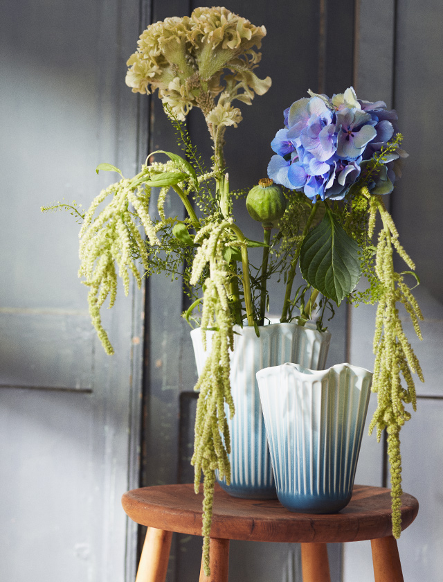 Stillleben mit Kähler Design Hammershøi Unfold Vasen in Blau: große und kleine Vase mit Farbverlauf, gefüllt mit Blumen und hängendem Grün auf einem Holzstuhl