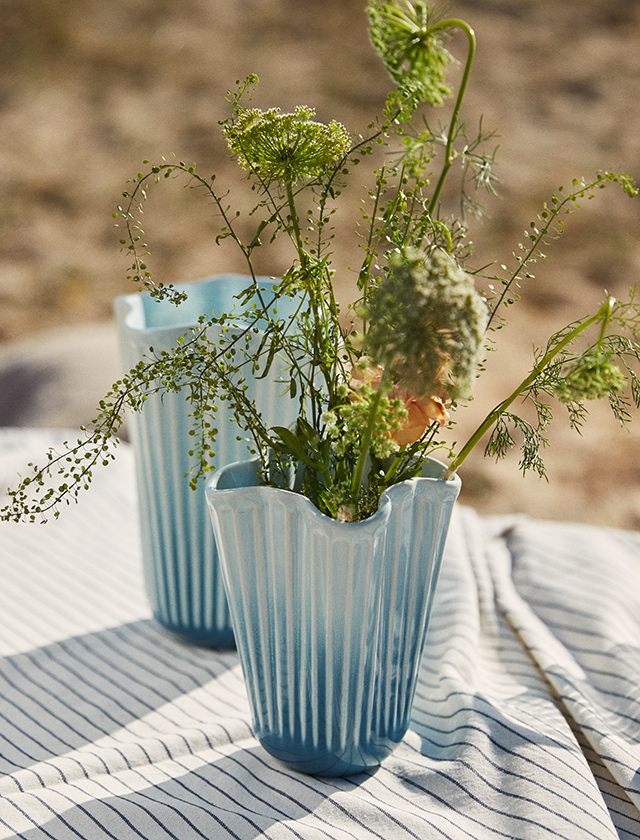 Sommerliches Stillleben: große und kleine Hammershøi Unfold Vase in Blau, gefüllt mit wilden grünen Stielen und Blüten, fotografiert im warmen Sonnenlicht am Strand