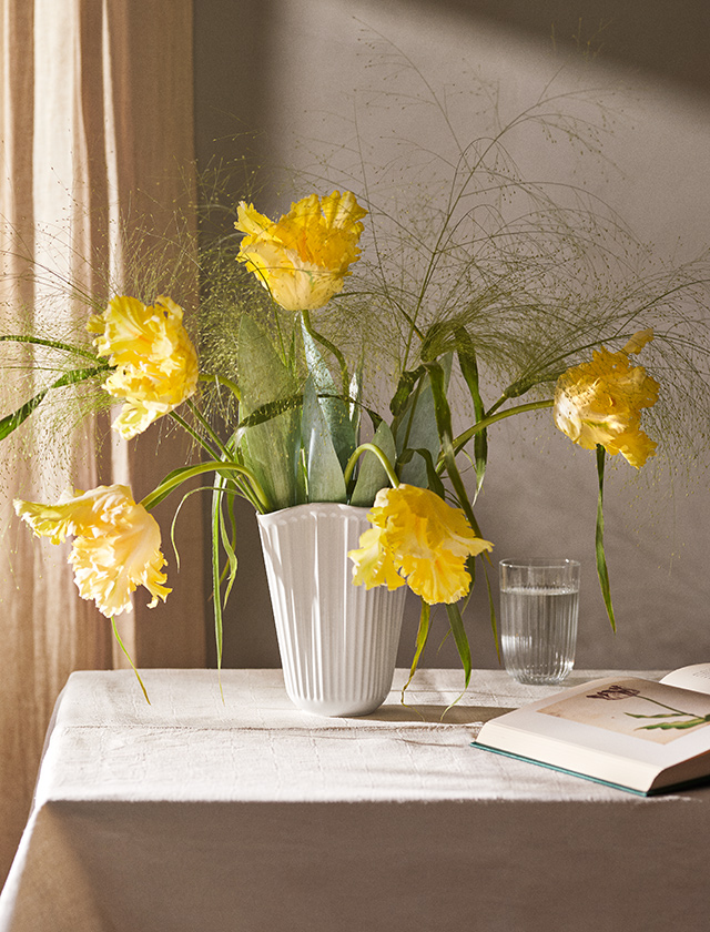 Weiße Kähler Hammershøi Unfold Vase aus Keramik mit gelben Tulpen und Ziergras, dekorativ auf einem Tisch neben einem Glas Wasser und einem geöffneten Buch – stilvolle skandinavische Wohnidee.