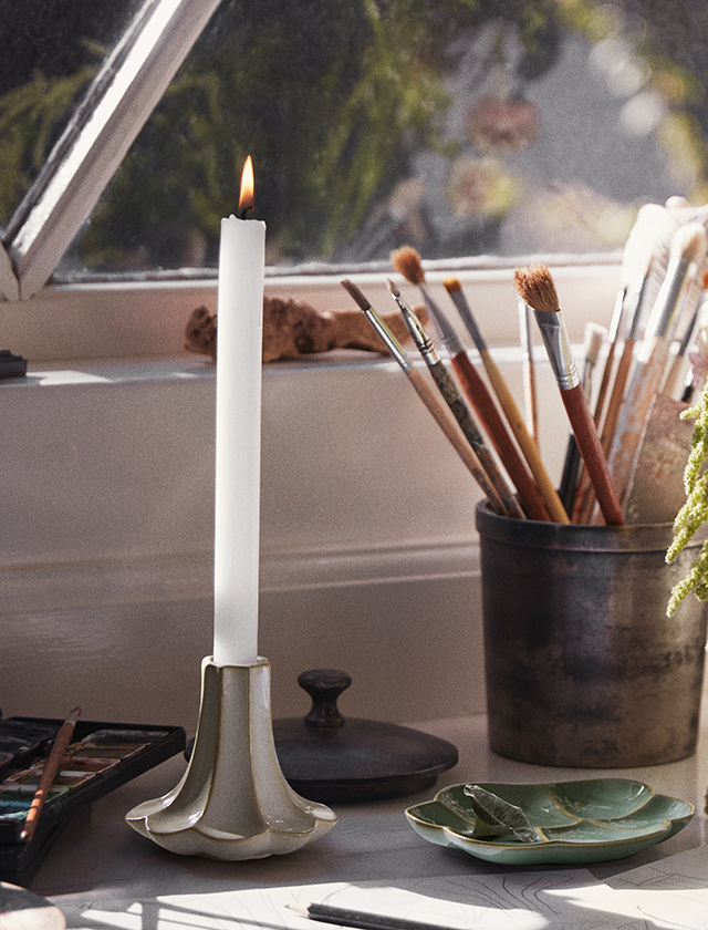 Brennende Stabkerze im weißen Kantarel Kerzenleuchter aus glasierter Keramik auf der Fensterbank, neben Pinseln und Aquarellkasten.