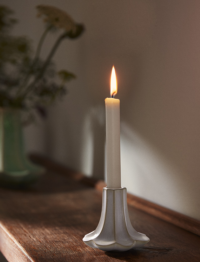 Weißer Kantarel Kerzenhalter aus Keramik mit brennender Stabkerze auf Holzoberfläche, Vase unscharf im Hintergrund.