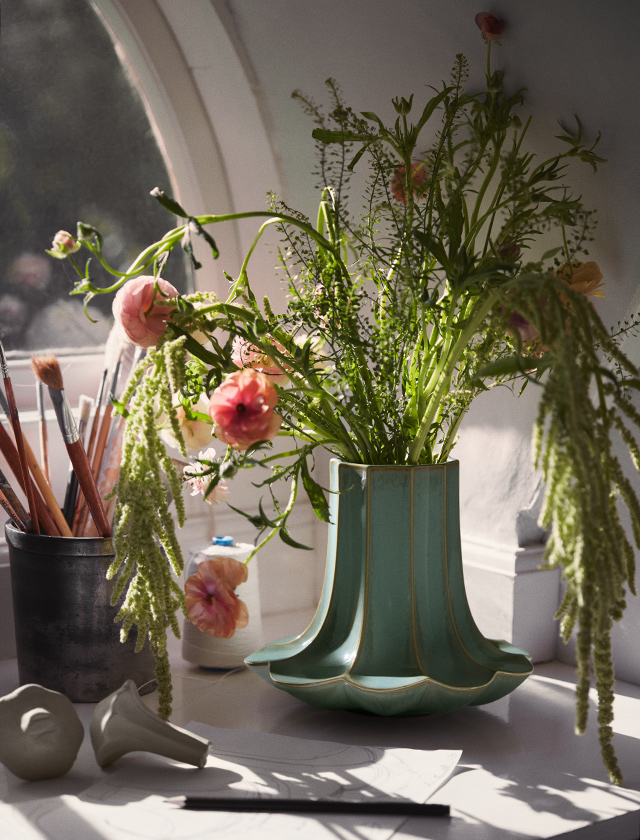 Grüne Kantarel Keramikvase mit lockerem Blumenstrauß auf der Fensterbank im warmen Tageslicht, mit Atelier-Details im Hintergrund.