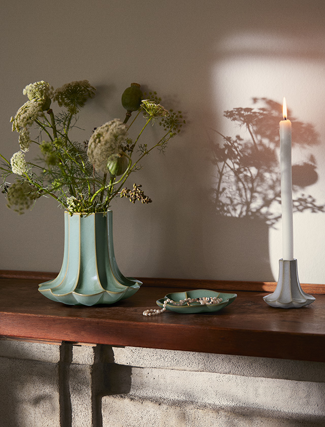 Mintgrüne Kantarel Vase mit Wildblumen, kleine Schale mit Perlen und Kerzenhalter mit weißer Kerze auf einer Holzablage im Sonnenlicht.
