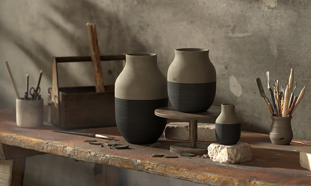 Wundervolles Ensemble von drei Vasen aus der Omaggio Circulare-Serie von Kähler Design, platziert in einer Töpferei. Die Vasen variieren in der Größe und präsentieren ein harmonisches zweifarbiges Design mit einer oberen Hälfte in einem natürlichen, erdigen Ton und einer unteren Hälfte, die in einem dunklen, matten Schwarz gehalten ist. Sie ruhen auf einer robusten Werkbank, die von handwerklichen Werkzeugen und Zubehör umgeben ist – darunter Pinsel, Spatel und ein rustikaler Metallkasten –, was die handwerkliche Umgebung betont.  Das sanfte Licht und die Schatten unterstreichen das handgemachte und künstlerische Flair der Szenerie.