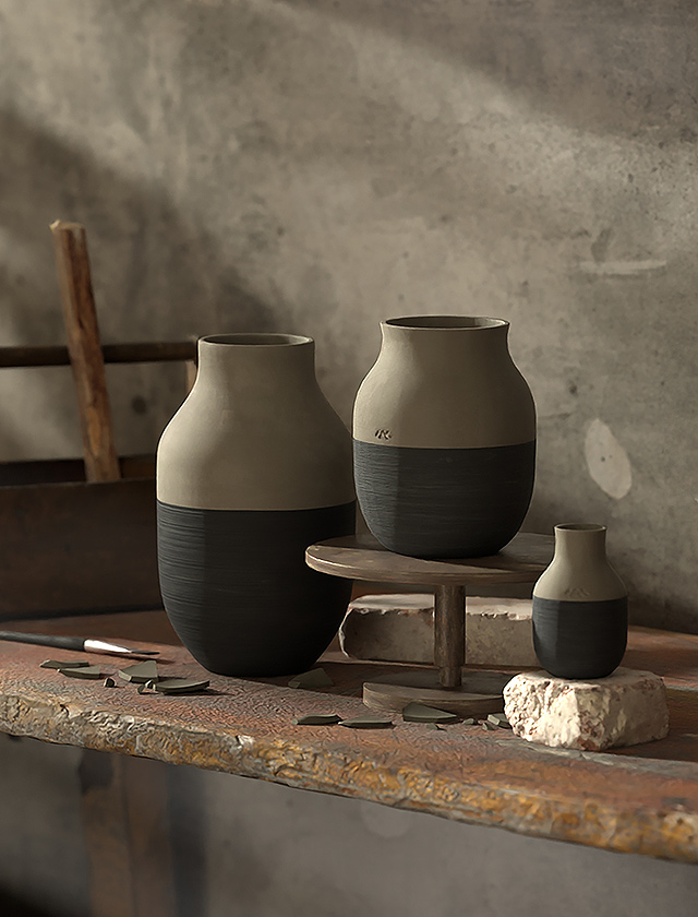 Das Bild zeigt einen näheren Blick auf drei Keramikvasen, die scheinbar Teil der Kaehler-Design-Omaggio-Circulare-Serie sind, unterschieden durch ihre Größe: groß, mittel und klein. Die Vordergrundvasen sind auf einer alten Holzoberfläche platziert, wobei die größere Vase auf einem kleinen, runden Holzpodest steht, was einen schönen Kontrast zum rustikalen Hintergrund bietet. Die Vasen zeichnen sich durch einen zweifarbigen Design aus: Der obere Teil ist in einem naturbelassenen Ton gehalten, während der untere Teil dunkelgrau ist. Im Hintergrund ist ein Teil einer Holzleiter und die gleiche unverputzte Betonwand wie im vorherigen Bild zu erkennen. Auf der Arbeitsfläche sind auch Tonfragmente und Werkzeuge verteilt, die auf die Handwerkskunst des Töpferns hinweisen. Das Bild fängt eine ruhige und künstlerische Atmosphäre ein und fokussiert sich auf die einfache, aber elegante Schönheit der Keramikvasen.