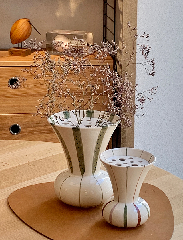 Signature Vasen in der großen und kleinen Größe mit Loch-Einsatz zum arrangieren von einzelnen Zweigen, Blumen und Stängeln von Kähler Design aus Dänemark - Arrangiert auf einem Björk Tisch von FDB Møbler, einem Platzset von LIND DNA. Im Hintergrung ist ein String Regal in Nussbaum von String Furnirture und ein Beak Bird von Warm Nordic