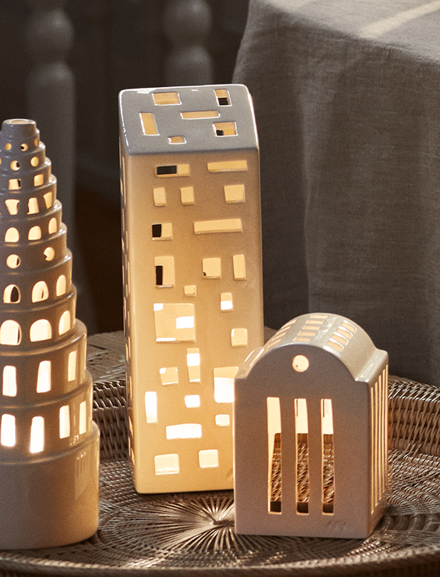 Das Kähler Design Urbania Teelichthaus „High Building“ aus weiß glasierter Keramik verbreitet ein warmes, gemütliches Licht. Die moderne Hochhaus-Silhouette mit unregelmäßig gesetzten Fensteröffnungen schafft ein faszinierendes Schattenspiel und bringt urbanes Flair ins Zuhause. In Kombination mit weiteren Urbania Lichthäusern entsteht eine kleine leuchtende Stadtlandschaft – ideal für Fensterbank, Tisch oder Sideboard.