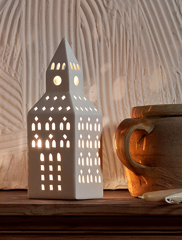 Das Urbania Kirchturm Lichthaus von Kähler als Einzelstück in Szene gesetzt. Durch die filigranen Fensteröffnungen strahlt das warme Kerzenlicht, das sich in der weißen Keramik reflektiert und ein elegantes Spiel aus Licht und Schatten erzeugt.