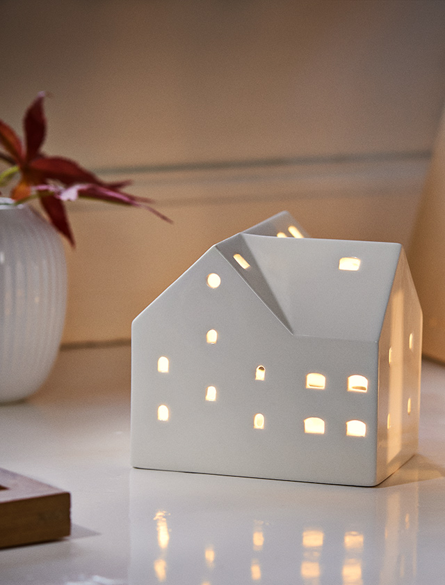 Kähler Design Urbania Teelichthaus Stall 2026 aus weißem Porzellan mit warm leuchtenden Fenstern – skandinavische Weihnachtsdekoration und stimmungsvolles Lichthaus.